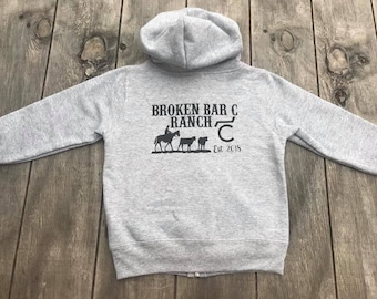 May include: Gray zip-up hoodie with a black graphic of a cowboy riding a horse and two cows. The graphic says "Broken Bar C Ranch Est. 2018".