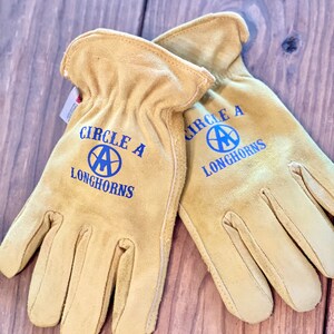 May include: A pair of tan leather work gloves with blue stitching and the text "Circle A Longhorns" on each glove.