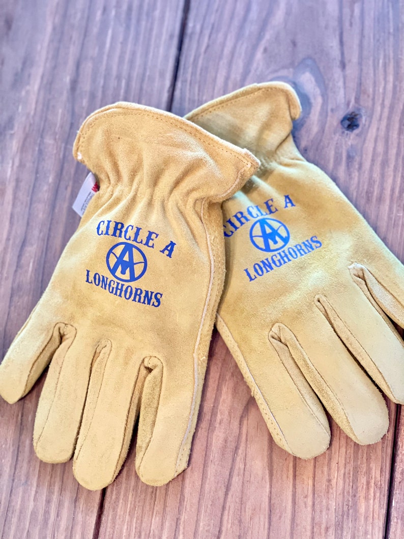 May include: A pair of tan leather work gloves with blue stitching and the text "Circle A Longhorns" on each glove.