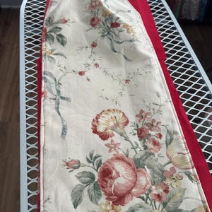May include: Cream-colored floral table runner with a red border. The runner features a repeating pattern of pink and red roses, carnations, and other flowers with green leaves and blue ribbons. The runner is placed on a white metal surface.