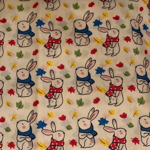 May include: A white fabric with a repeating pattern of white bunnies wearing scarves. The bunnies are wearing blue and red scarves with white polka dots. The background is a cream color with yellow, red, and green leaves.