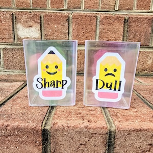 May include: Two clear plastic containers with pencil-themed designs. One features a smiling pencil with the word "Sharp," and the other has a sad pencil with the word "Dull." The containers are set against a brick background.