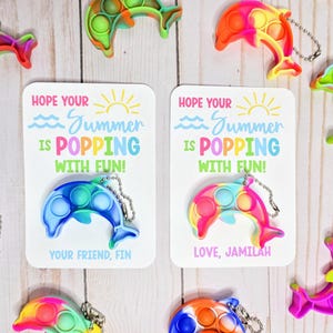 May include: Colorful dolphin-shaped pop-it keychains in various colors, including blue, pink, orange, and green. Each keychain has a metal chain. Two cards read "Hope your Summer is POPPING with FUN!" One card says "Your friend, Fin" and the other says "Love, Jamilah."