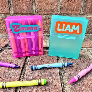 May include: Two plastic crayon boxes, one pink and one turquoise, each labelled with a name. The pink box says "Emma" in a teal font, and the turquoise box says "Liam" in an orange font. Several crayons are scattered around the boxes.