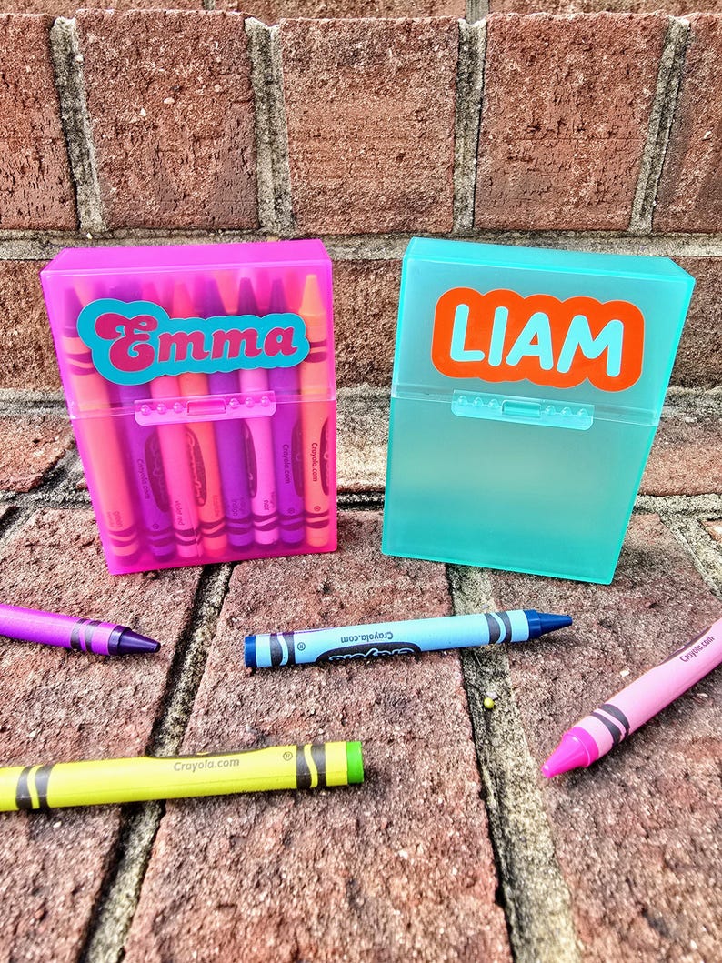 May include: Two plastic crayon boxes, one pink and one turquoise, each labelled with a name. The pink box says "Emma" in a teal font, and the turquoise box says "Liam" in an orange font. Several crayons are scattered around the boxes.