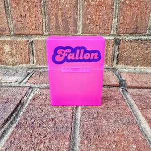 May include: A vibrant pink plastic box, featuring the name "Fallon" in a retro style. The box is rectangular with a hinged lid and a small clasp. The background is a brick wall.