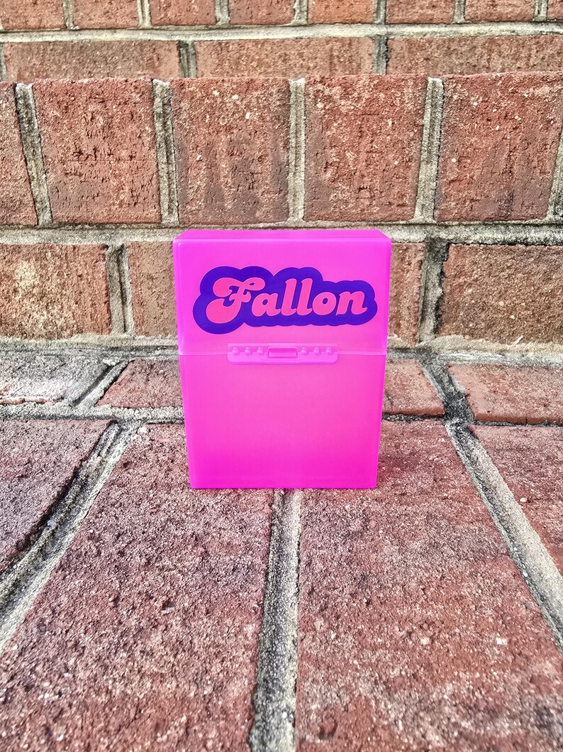 May include: A vibrant pink plastic box, featuring the name "Fallon" in a retro style. The box is rectangular with a hinged lid and a small clasp. The background is a brick wall.