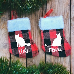 May include: Two red and black buffalo plaid Christmas stockings with white cat silhouettes and the names "Loki" and "Oliver" written below.