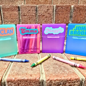 May include: Four colourful crayon boxes, each labelled with a name: Declan, Emma, Maggie, and John Brooks. The boxes are teal, pink, purple, and blue. Crayons in various colours are scattered in front of the boxes.