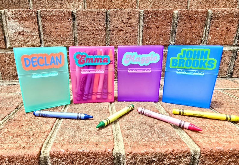 May include: Four colourful crayon boxes, each labelled with a name: Declan, Emma, Maggie, and John Brooks. The boxes are teal, pink, purple, and blue. Crayons in various colours are scattered in front of the boxes.