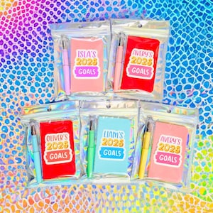 May include: A collection of small notebooks and pens in clear plastic packaging. The notebooks are in various colors, including pink, red, and blue, with the text "2025 GOALS" and names like "Isla's" and "Avery's". The pens match the notebook colors.