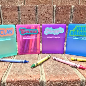 May include: Four coloured plastic crayon boxes, each labelled with a name: Declan, Emma, Maggie, and John Brooks. Each box is a different colour: teal, pink, purple, and blue. Crayons of various colours are scattered around the boxes.