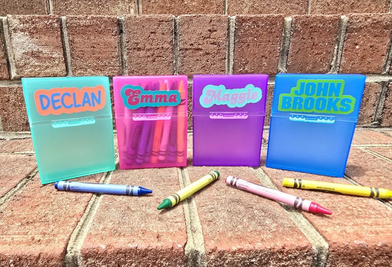 May include: Four coloured plastic crayon boxes, each labelled with a name: Declan, Emma, Maggie, and John Brooks. Each box is a different colour: teal, pink, purple, and blue. Crayons of various colours are scattered around the boxes.