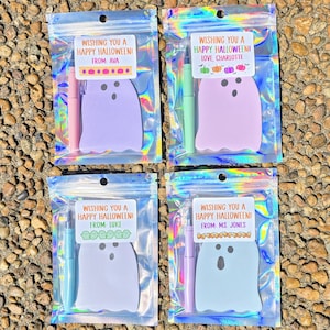 May include: Four Halloween-themed gift sets in iridescent bags. Each set includes a ghost-shaped notepad and a pen. The labels read "Wishing You a Happy Halloween!" with different names: Ava, Charlotte, Luke, and Ms. Jones. The notepads and pens are in various pastel colors.