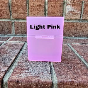 May include: A light pink, rectangular box with a snap closure. The text "Light Pink" is printed in black on the front of the box. The box is on a brick surface.