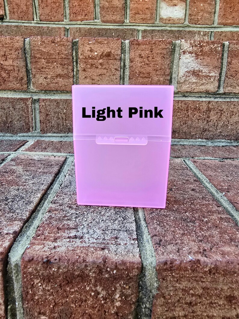 May include: A light pink, rectangular box with a snap closure. The text "Light Pink" is printed in black on the front of the box. The box is on a brick surface.