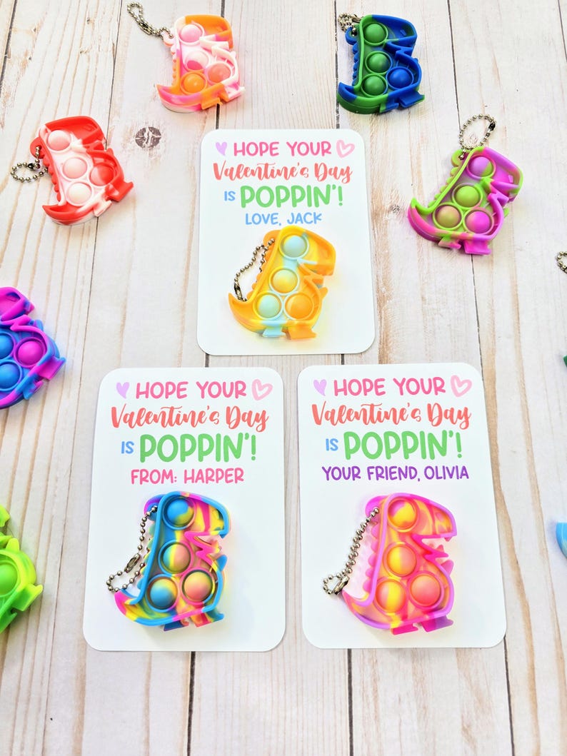 May include: Three Valentine's Day cards with colorful dinosaur-shaped pop-it fidget toys attached. The cards have the text "Hope your Valentine's Day is Poppin'!". One card says "Love, Jack", another says "From: Harper", and the third says "Your friend, Olivia".