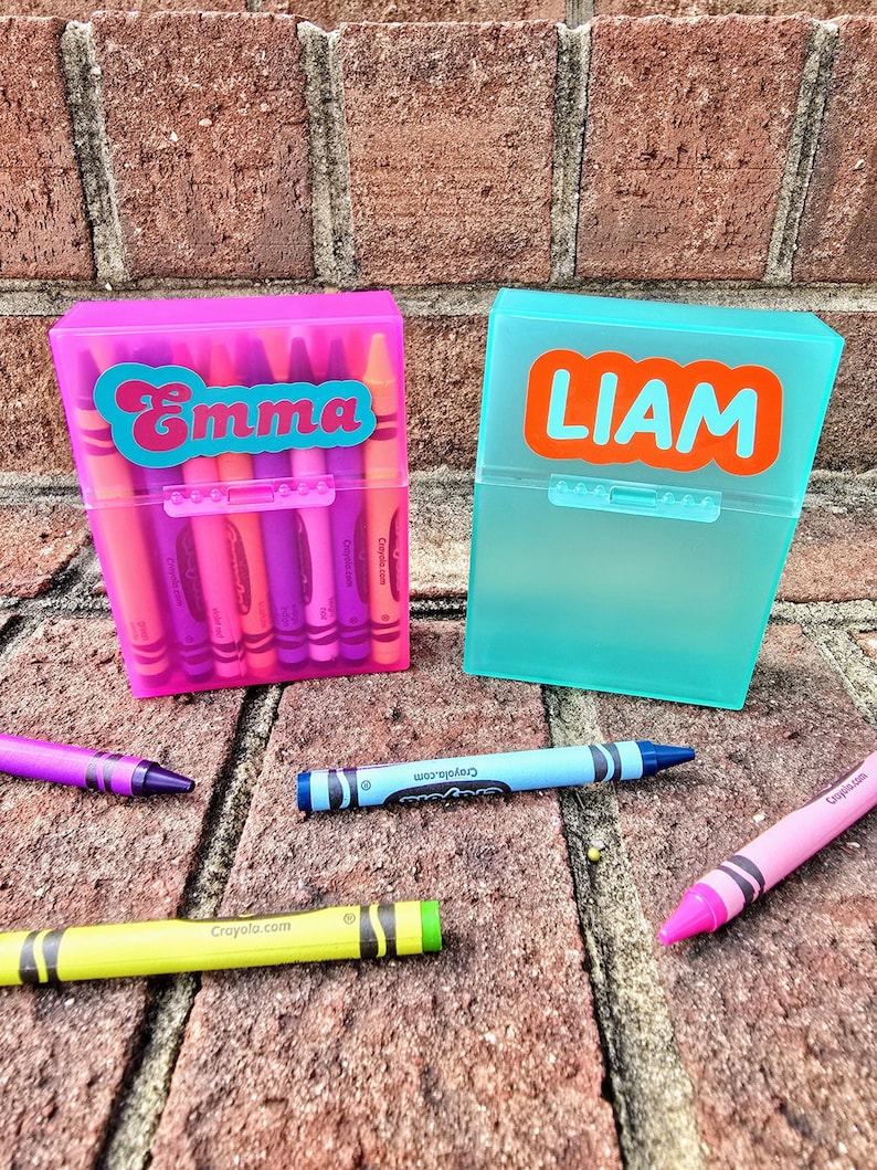 May include: Two plastic crayon boxes, one pink and one teal, each labelled with a name. The pink box says "Emma" in a teal font, and the teal box says "LIAM" in an orange font. Several crayons are scattered around the boxes.