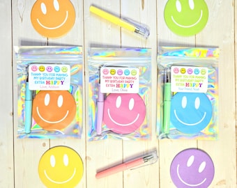 May include: Three party favor bags with smiley face stickers and pens. Each bag contains a smiley face sticker in orange, pink, and blue, and a pen in a matching color. The bags have a label that says "THANK YOU FOR MAKING MY BIRTHDAY PARTY EXTRA HAPPY."