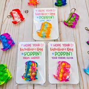 May include: A set of Valentine's Day cards featuring colorful dinosaur-shaped pop-it toys. Each card has a message that says "Hope your Valentine's Day is poppin'!". The cards are ready to be handed out.