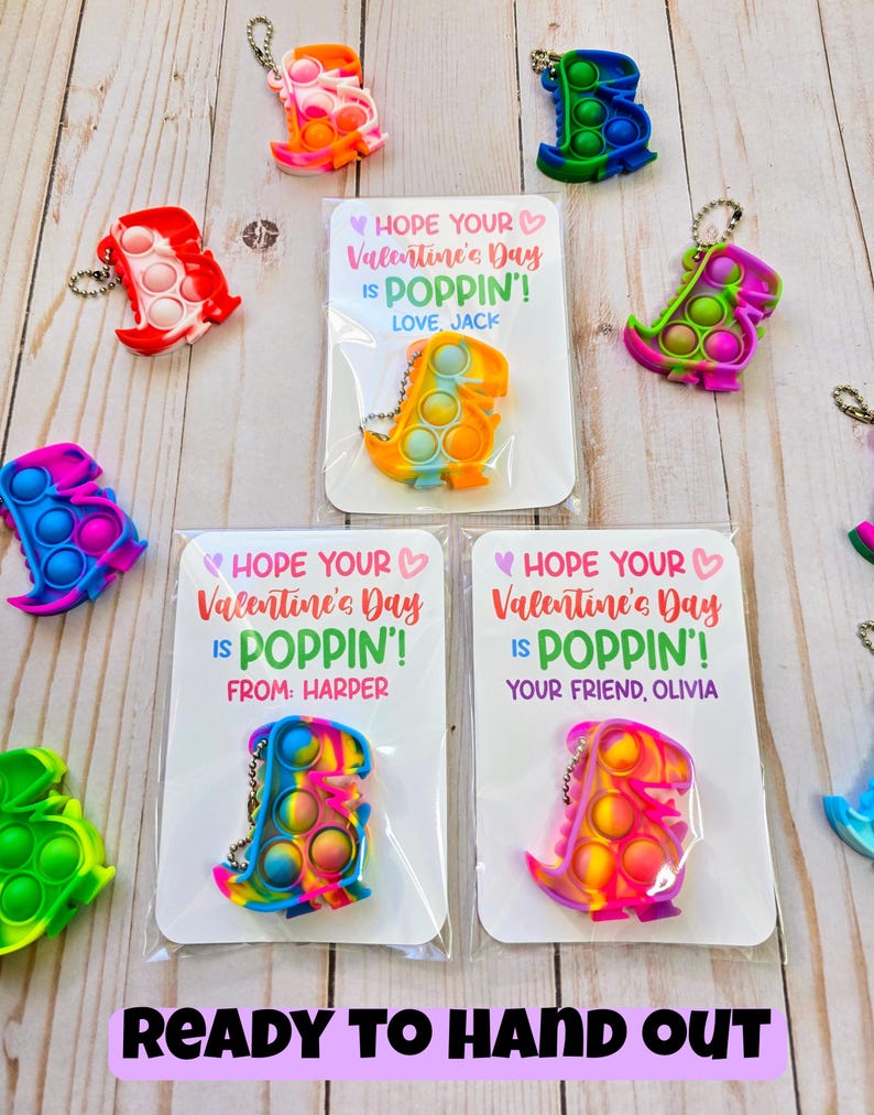 May include: A set of Valentine's Day cards featuring colorful dinosaur-shaped pop-it toys. Each card has a message that says "Hope your Valentine's Day is poppin'!". The cards are ready to be handed out.