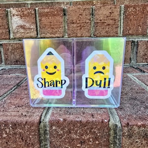 May include: A clear, divided container with iridescent sides. Each section has a pencil-shaped label. One label reads "Sharp" with a smiling pencil, the other reads "Dull" with a frowning pencil. The container sits on a brick surface.