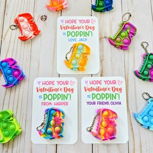 May include: Valentine's Day cards with a pop-it dinosaur keychain attached. The cards have a pink heart and the text "Hope your Valentine's Day is poppin'!". The cards are personalized with names like "Jack", "Harper", and "Olivia".