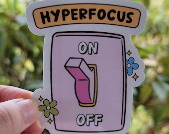 May include: A sticker featuring a pink switch design with the word "OFF" in black text. The sticker has the words "HYPERFOCUS" in black text. The sticker is decorated with cartoon flowers. The switch is in the "off" position.