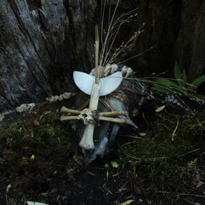 May include: A handcrafted bone and wood sculpture with a cross-like structure, adorned with a white, wing-shaped element and dried grasses. The sculpture is set against a backdrop of moss and a tree trunk, creating a natural, earthy aesthetic.