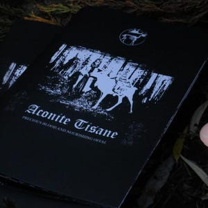 May include: Two black brochures with white text and illustrations. The brochures feature the text "Aconite Tisane" and an illustration of a deer. A lit white candle is in the background.