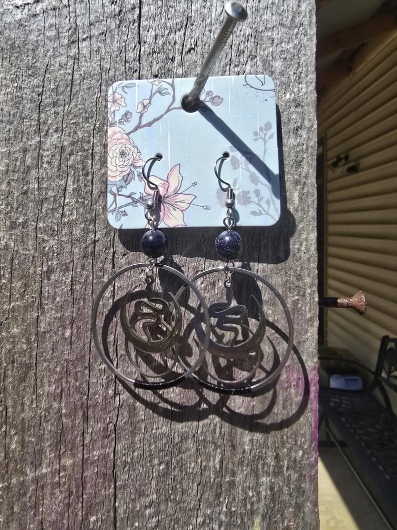 Puede incluir: Un par de pendientes de aro de plata con una cuenta de piedra azul en cada pendiente. Los pendientes cuelgan de una tarjeta con estampado floral.