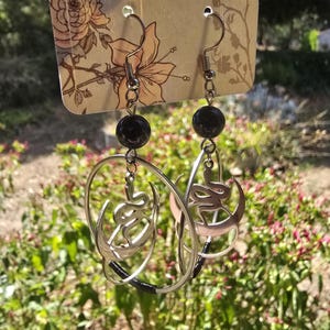 Puede incluir: Un par de pendientes de aro de plata con una cuenta negra en la parte superior. Cada aro presenta una luna creciente de plata con un dise&ntilde;o de serpiente en su interior.