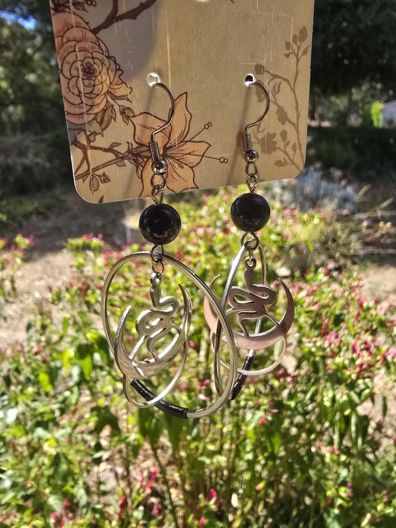 Puede incluir: Un par de pendientes de aro de plata con una cuenta negra en la parte superior. Cada aro presenta una luna creciente de plata con un dise&ntilde;o de serpiente en su interior.
