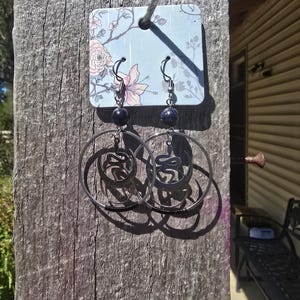 Peut inclure: Une paire de boucles d'oreilles créoles noires avec un motif de serpent. Les boucles d'oreilles sont suspendues à une carte bleue à motif floral. La carte est fixée à une surface en bois.