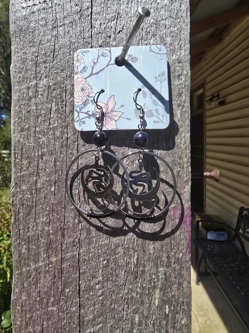 Puede incluir: Un par de pendientes de aro negros con un dise&ntilde;o de serpiente. Los pendientes cuelgan de una tarjeta azul con un patr&oacute;n floral. La tarjeta est&aacute; unida a una superficie de madera.