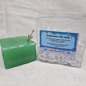 May include: A green gelatinous cube soap with a silver miniature sword charm. The soap is packaged in a clear plastic box with white shredded paper. The label on the box reads "Gelatinous Cube Soap" and "You wish dungeon dwellers smelled this nice! Each gelatinous cube contains a mini 7-piece dice set." The website address "Shellerbellesoapary.etsy.com" is printed at the bottom of the label.