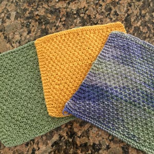 May include: Three knitted dishcloths in green, yellow, and purple. The dishcloths are made with a simple stitch pattern and are ready to be used for cleaning.