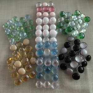 May include: Assortment of glass mosaic tiles in various colors, including green, blue, white, pink, gold, and black. The tiles are arranged in a decorative pattern.