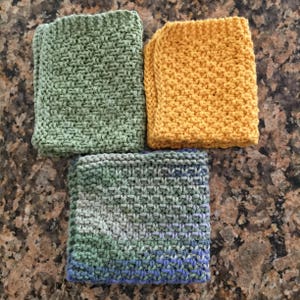 May include: Three knitted dishcloths in different colors: green, yellow, and a multi-colored green, blue, and gray. The dishcloths are made with a simple stitch pattern.