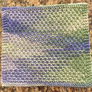 May include: A knitted dishcloth in shades of green, purple, and white. The dishcloth is made with a simple stitch pattern and has a slightly textured surface.