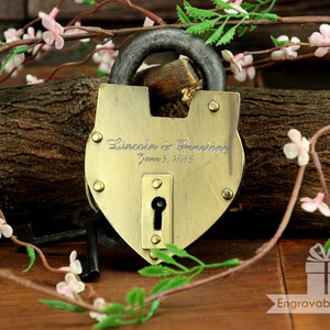 Puede incluir: Un candado de latón en forma de corazón con un ojo de cerradura y el texto grabado: "Lincoln & Penelope June 3, 2015". El candado está sobre una superficie de madera con flores rosas.