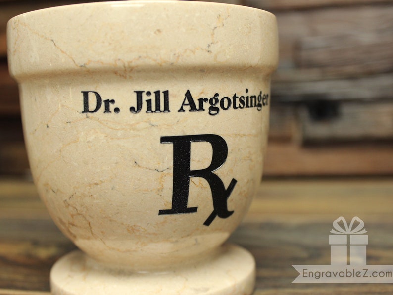 May include: A beige marble mortar and pestle with the name "Dr. Jill Argotsinger" and a large "Rx" symbol in black lettering. The mortar has a wide top and a rounded base.