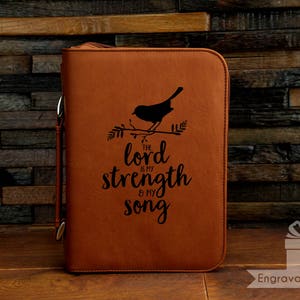 May include: A brown leather Bible cover with a black bird perched on a branch, and the words "The Lord is my strength & my song" in a script font. The cover has a zipper closure and a silver metal ring.