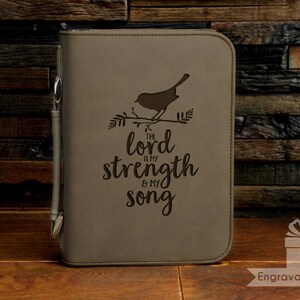 May include: A tan Bible cover with a bird and branch design, and the text "The Lord is my strength & my song." A pen is attached to the side. The cover is on a wooden surface.