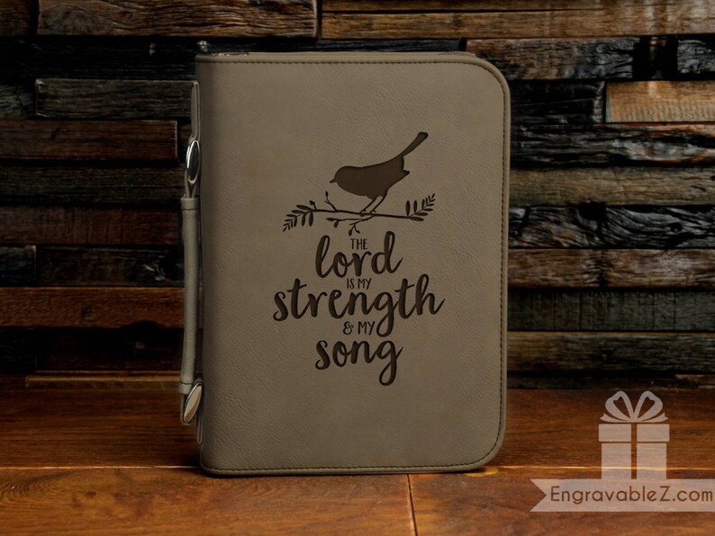 May include: A tan Bible cover with a bird and branch design, and the text "The Lord is my strength & my song." A pen is attached to the side. The cover is on a wooden surface.