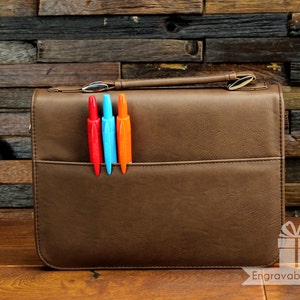 May include: Brown leather briefcase with a handle and a front pocket. Three colorful pens, red, blue, and orange, are in the pocket. The briefcase has a silver clasp and the text "EngravableZ.com" is visible.