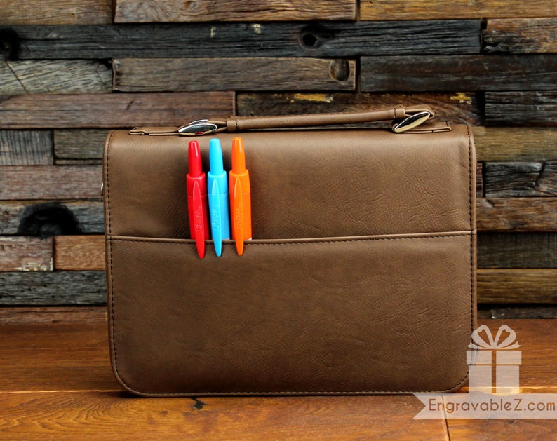 May include: Brown leather briefcase with a handle and a front pocket. Three colorful pens, red, blue, and orange, are in the pocket. The briefcase has a silver clasp and the text "EngravableZ.com" is visible.