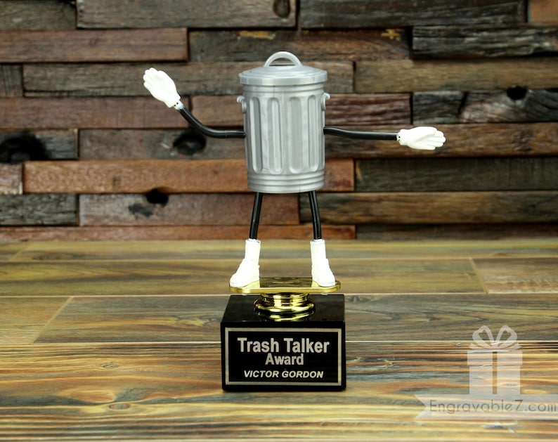 May include: A silver trash can with arms and legs, wearing white gloves and boots, stands on a black base with a gold plaque that reads "Trash Talker Award VICTOR GORDON".