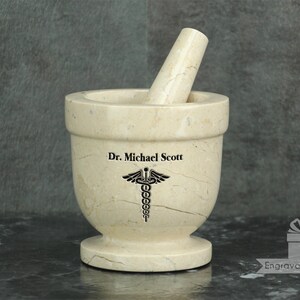 May include: A beige marble mortar and pestle set. The mortar has a wide bowl and a footed base. The pestle rests inside the bowl. The text "Dr. Michael Scott" and a medical symbol are etched on the mortar.