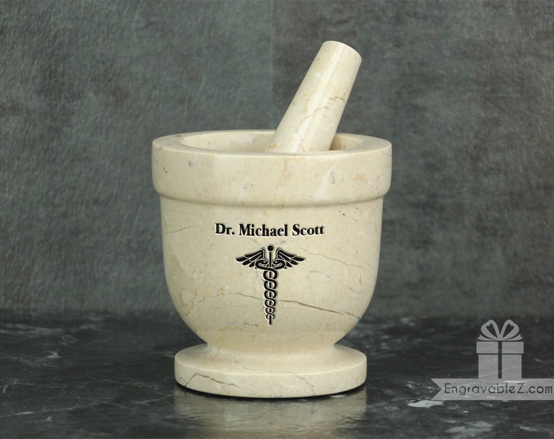 May include: A beige marble mortar and pestle set. The mortar has a wide bowl and a footed base. The pestle rests inside the bowl. The text "Dr. Michael Scott" and a medical symbol are etched on the mortar.